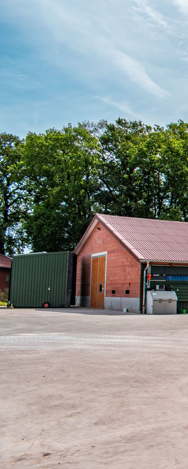 Zwei rote Backsteingebäude mit großen Holztüren, umgeben von einem gepflasterten Hof und Bäumen im Hintergrund.