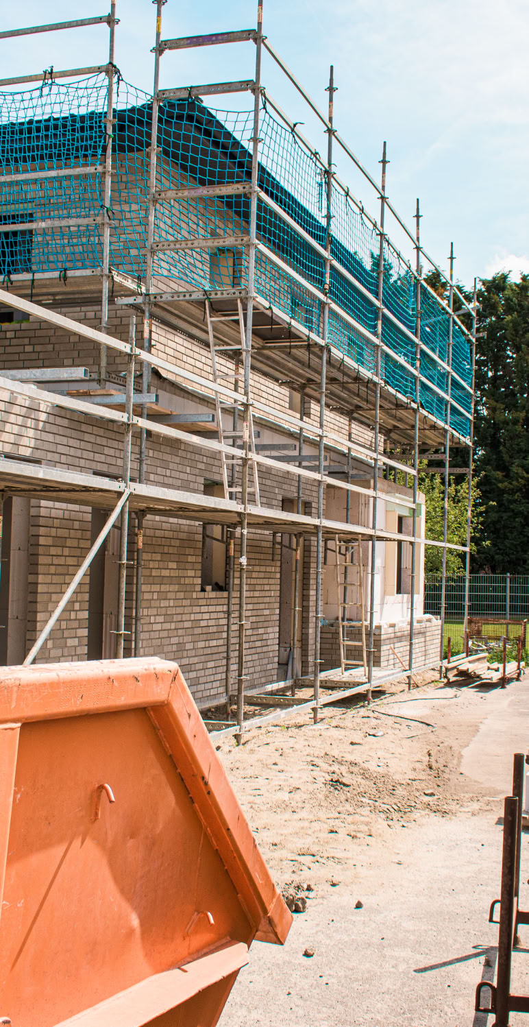 Ein Gebäude im Bau mit Gerüsten und Baumaterialien, im Vordergrund ein orangefarbener Container, im Hintergrund Bäume.