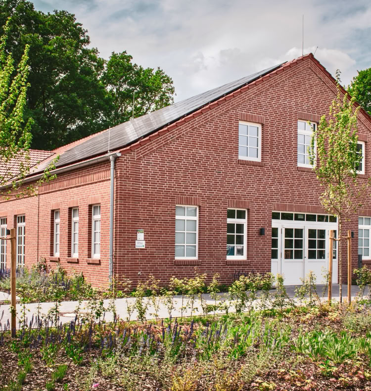 Backsteingebäude mit weißen Fenstern und Türen, umgeben von Bäumen und Pflanzen.