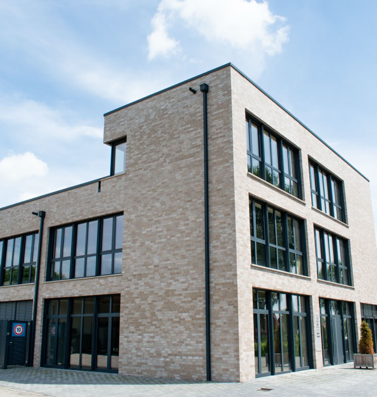 Modernes, dreistöckiges Gebäude aus Backstein mit großen Fenstern, umgeben von einem gepflasterten Bereich und Bäumen im Hintergrund.