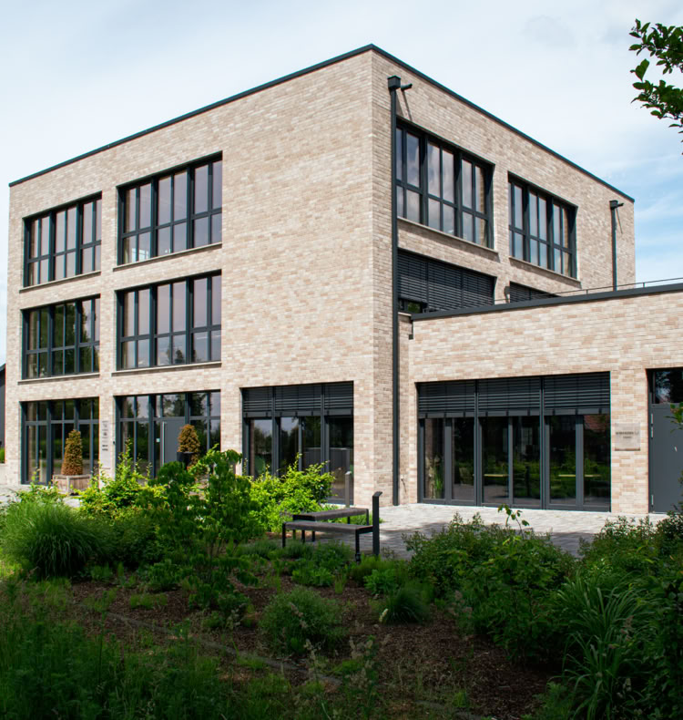 Modernes Gebäude aus Backstein mit großen Fenstern, umgeben von grüner Vegetation und einem Fahnenmast links.