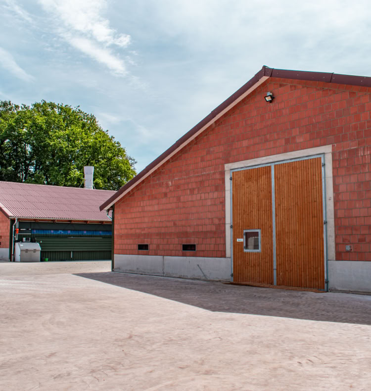 Zwei rote Backsteingebäude mit Holztüren auf einem Hof, im Hintergrund Bäume und ein blauer Himmel.