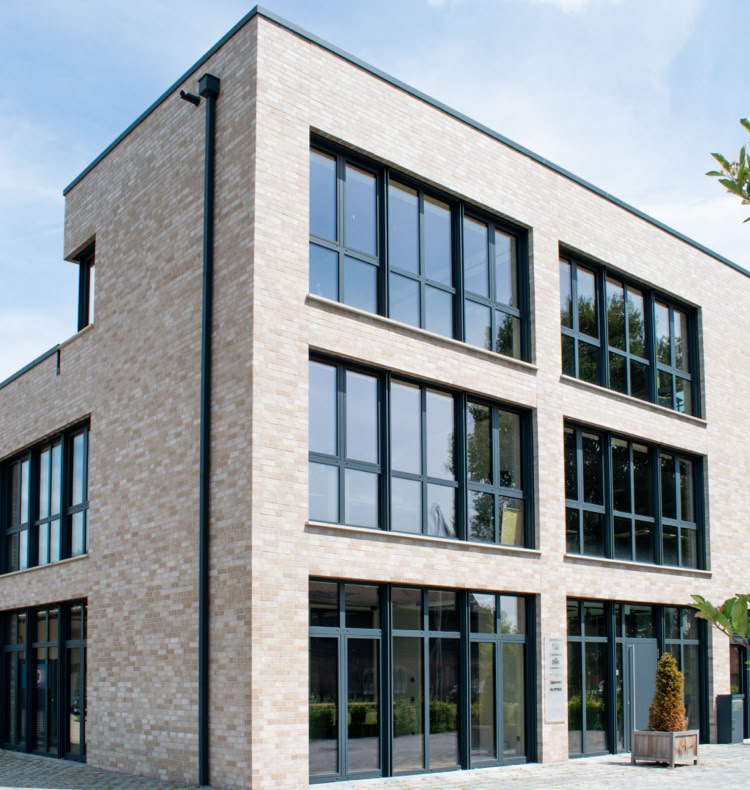 Modernes dreistöckiges Gebäude mit großen Fenstern und heller Backsteinfassade, umgeben von einem klaren Himmel und einigen Bäumen.