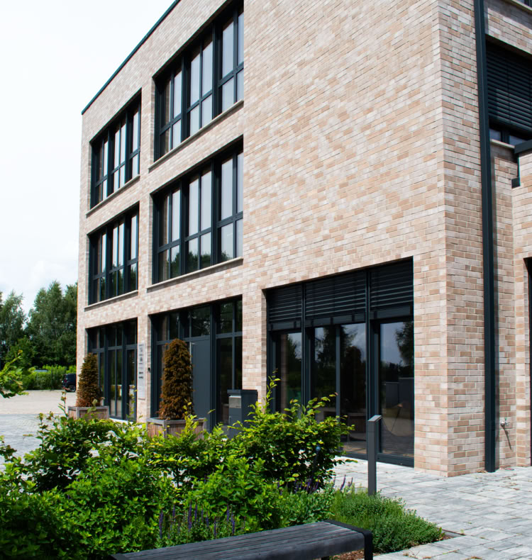 Backstein-Gebäude mit großen Fenstern, umgeben von Pflanzen und einem Gehweg, im Vordergrund eine Bank.