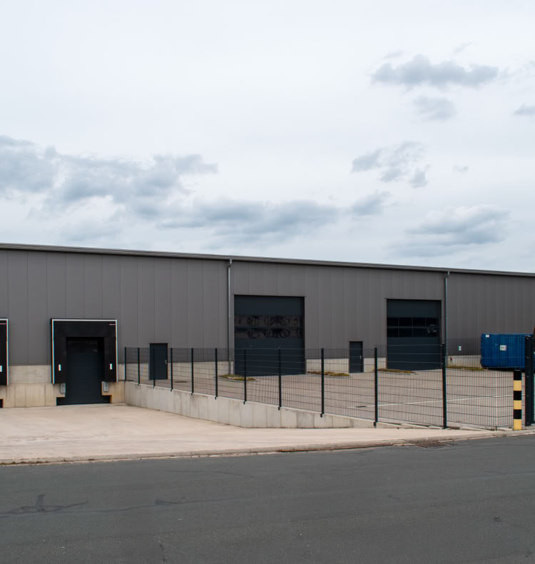 Grau gestrichenes Industriegebäude mit mehreren Laderampen und einem umzäunten Bereich, davor eine asphaltierte Straße.