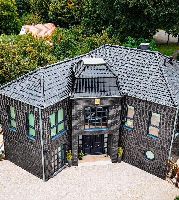 Großes, modernes Herremhaus mit dunkler Fassade und grauem Dach, umgeben von grünen Bäumen und Rasenflächen.