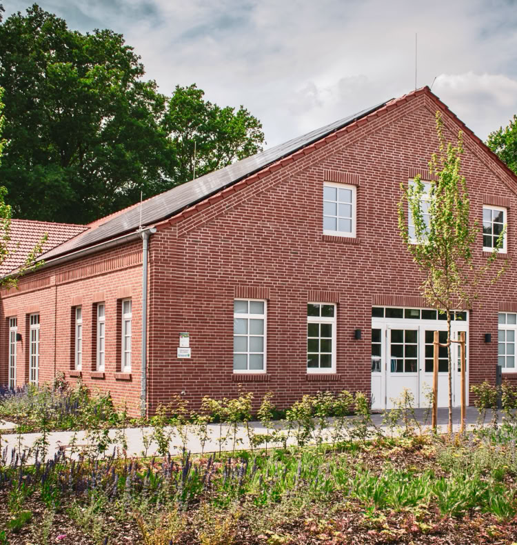 Backsteingebäude mit weißen Fenstern und Türen, umgeben von Bäumen und einem Garten im Vordergrund.