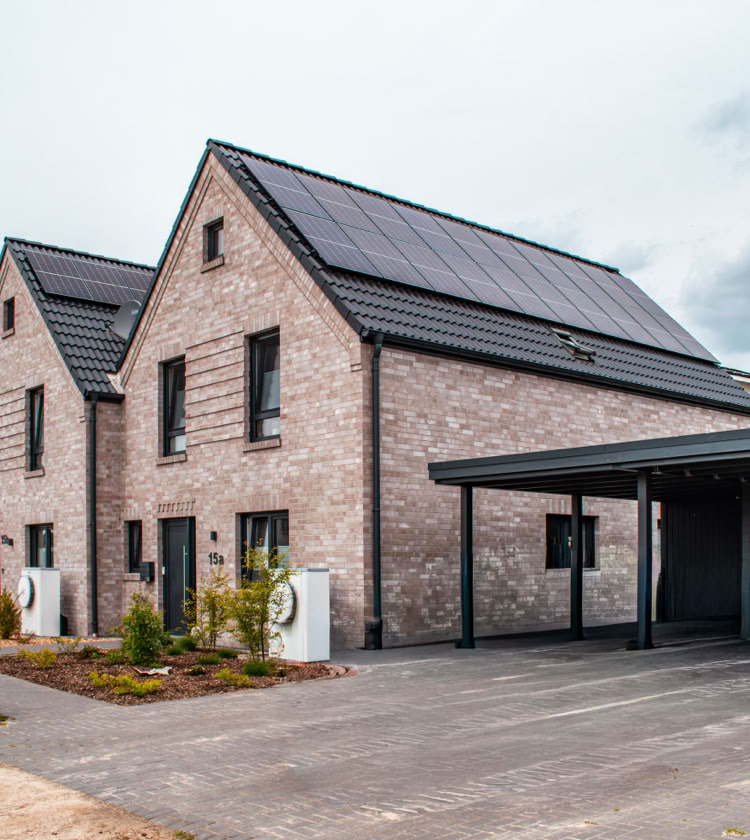 Reihenhäuser mit Ziegeldächern und Solarpaneelen, daneben ein Carport, umgeben von einem gepflasterten Weg und etwas Vegetation.
