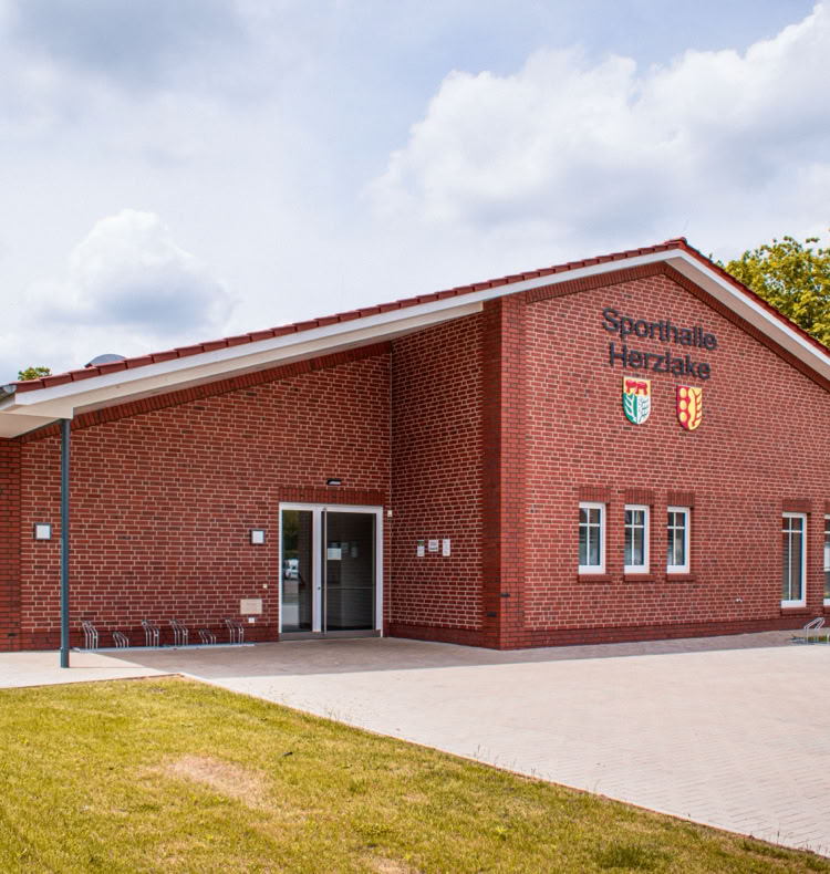 Backsteingebäude mit mehreren Fenstern und zwei Wappen an der Fassade, umgeben von einer Grünfläche und Bäumen, unter einem bewölkten Himmel.