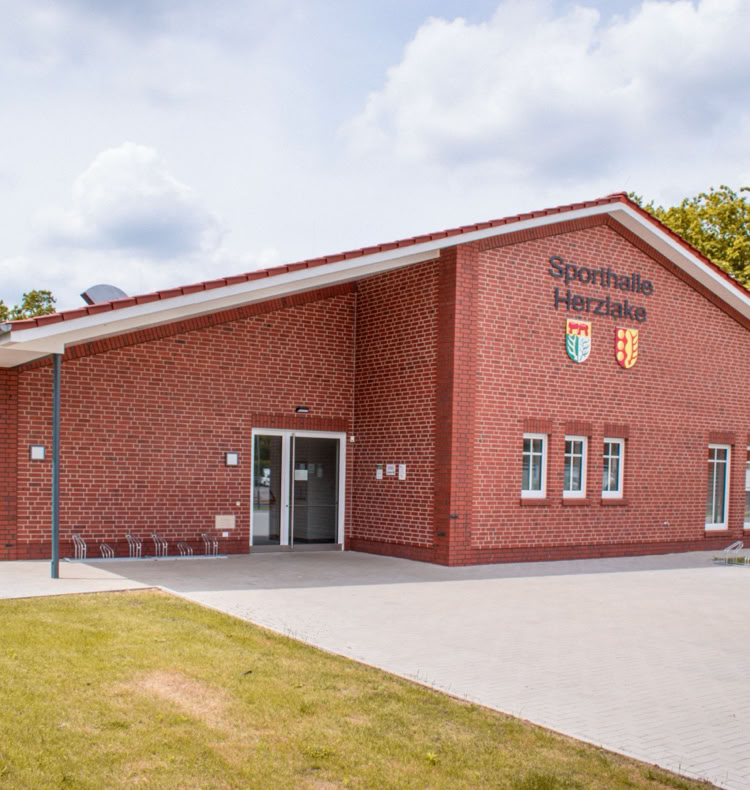 Backsteingebäude mit mehreren Fenstern und einem Eingang, umgeben von Rasen und Bäumen, unter einem bewölkten Himmel.